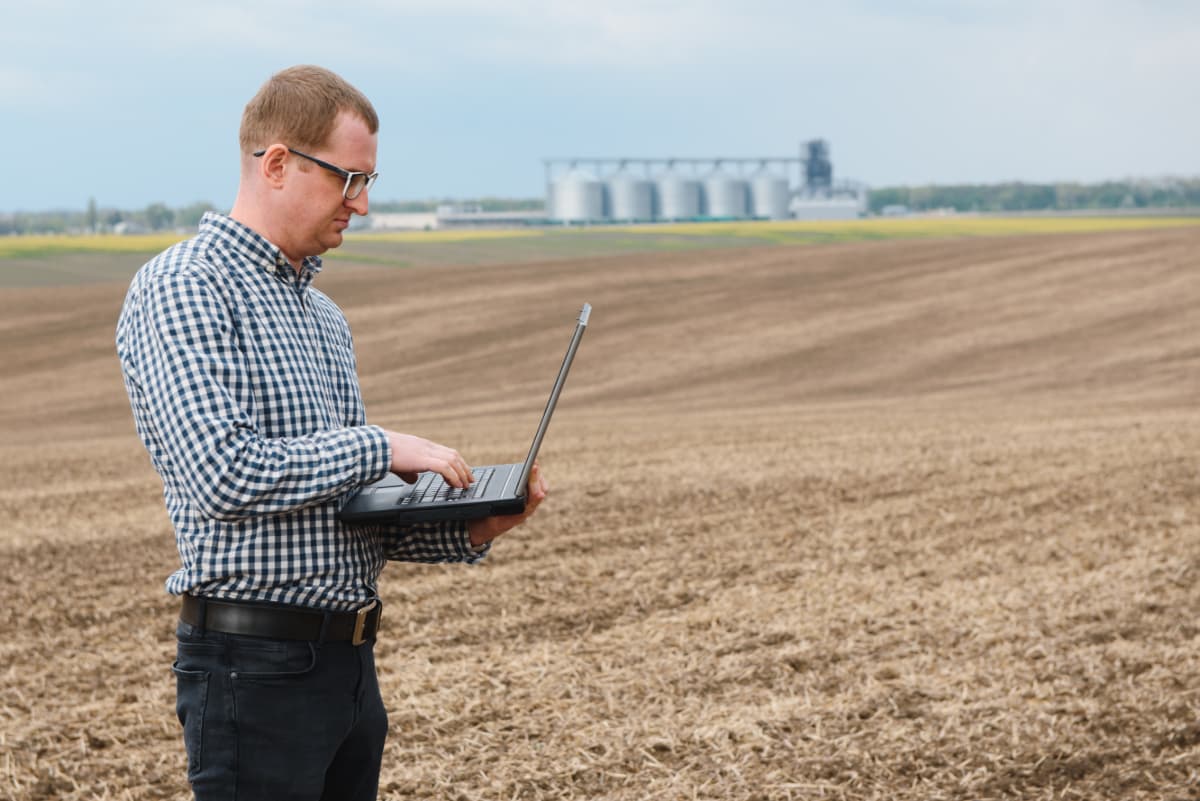 farmer-with-laptop-moisture-management A farmer using a laptop in a field. Grain bins are in the background.